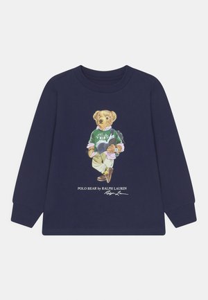 Chemise bleu marine à manches longues avec un ours portant un pull vert, un pantalon kaki et un sac à dos lisant un livre, étiquetée « Polo Bear par Ralph Lauren. »