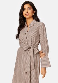 Abito a righe in marrone e crema, con vita accentuata da una cintura, maniche lunghe e polsini svasati, realizzato in un tessuto leggero.