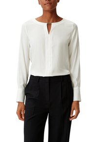 Femme portant un chemisier blanc à manches longues avec une petite ouverture en forme de goutte à l'avant et un pantalon noir ajusté, sur fond blanc.