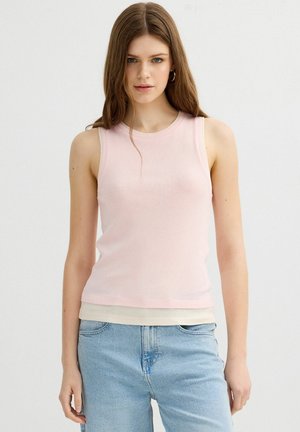 Joven mujer con cabello largo y castaño que lleva una camiseta sin mangas rosa claro y vaqueros de mezclilla azul claro sobre un fondo liso.