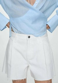 Shorts en denim blanc avec un ourlet effiloché, présentant des plis sur le devant et un détail de bouton noir à la taille. Portés avec une chemise bleu clair.