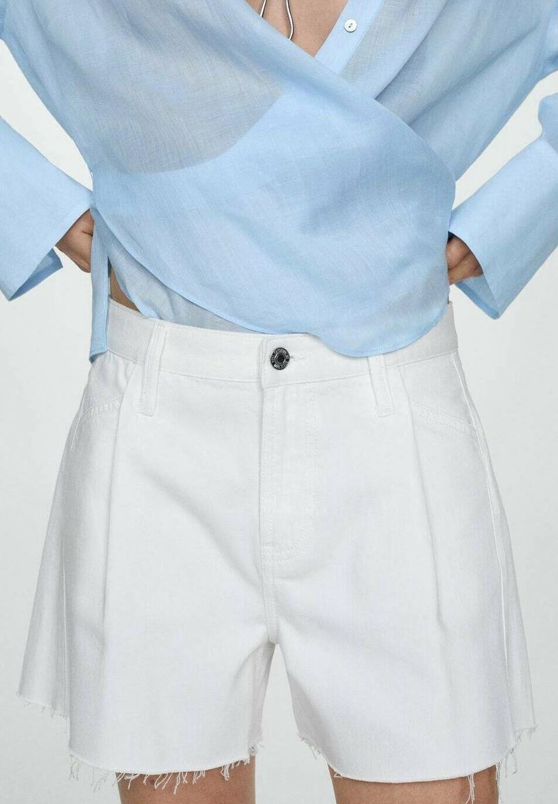 Shorts en denim blanc avec un ourlet effiloché, présentant des plis sur le devant et un détail de bouton noir à la taille. Portés avec une chemise bleu clair.