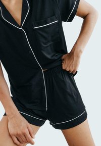 Czarny satynowy zestaw piżamowy z białym wykończeniem, składający się z krótkiego rękawa koszuli zapinanej na guziki oraz pasujących szortów z elastycznym pasem.