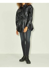 Musta nahkahiha takki, jossa on etunapit, kaksi rinta-taskua ja solmittava vyö, yhdistettynä tummiin farkkuihin ja mustiin nauhakenkiin.