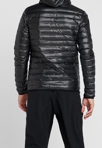 Veste matelassée noire avec une coupe ajustée, une texture matelassée horizontale et une capuche. Le matériau semble brillant et lisse.