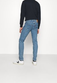 Osoba v lehce modrých slim-fit džínách, černé dlouhé tričko a bílé tenisky stojí proti bílé zdi, otočená zády.