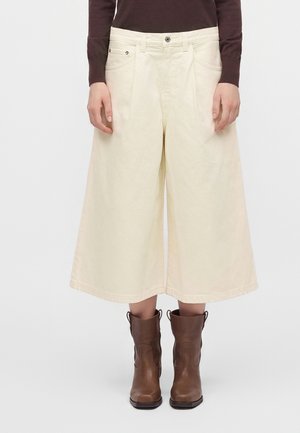 Personne portant un pantalon large crème coupé court, un haut à manches longues foncé et des bottes en cuir marron mi-mollet, debout devant un fond uni.