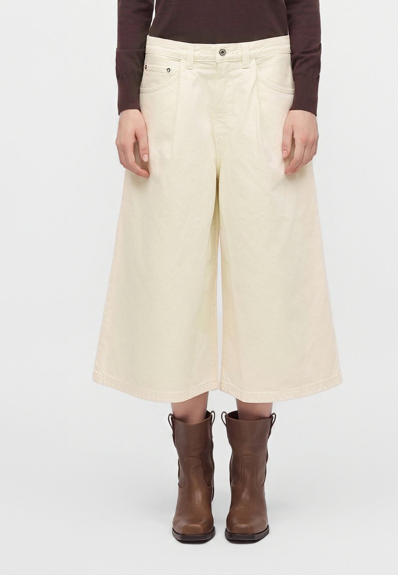 Personne portant un pantalon large crème coupé court, un haut à manches longues foncé et des bottes en cuir marron mi-mollet, debout devant un fond uni.
