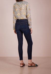 Blusa de manga larga con estampado floral y detalles dorados, combinada con jeans ajustados de mezclilla oscura y sandalias marrones con tiras.