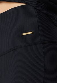 Tessuto attivo nero a coste con un badge metallico dorato inciso con la scritta "ELLEN NEW YORK." Texture liscia e design aderente.