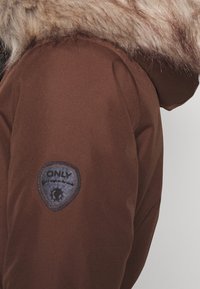 Hnědý rukáv zimní bundy s kožešinovou lemovkou kapuce a tmavou nášivkou s nápisem "ONLY" na horní části rukávu na jednobarevném pozadí.