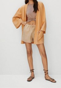 Lichtoranje kimono-stijl buitenkleding met wijde mouwen, gecombineerd met een bruin, getailleerd topje en beige shorts. Afgemaakt met bruin, verstelbare sandalen.