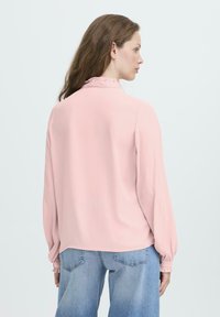Vrouw met lang bruin haar, gekleed in een lichtroze blouse met een gerimpelde kraag en manchetten, gecombineerd met lichtblauwe spijkerbroek, zijwaarts kijkend.