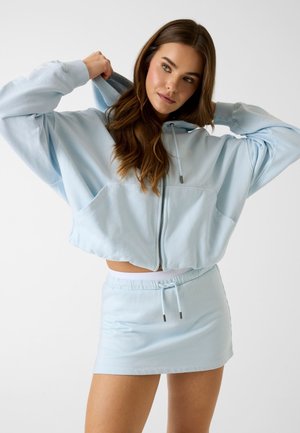 Frau, die die Kapuze eines hellblauen, kurz geschnittenen Kapuzenpullovers mit Reißverschluss anpasst, kombiniert mit einem passenden Minirock mit Kordelzug vor weißem Hintergrund.
