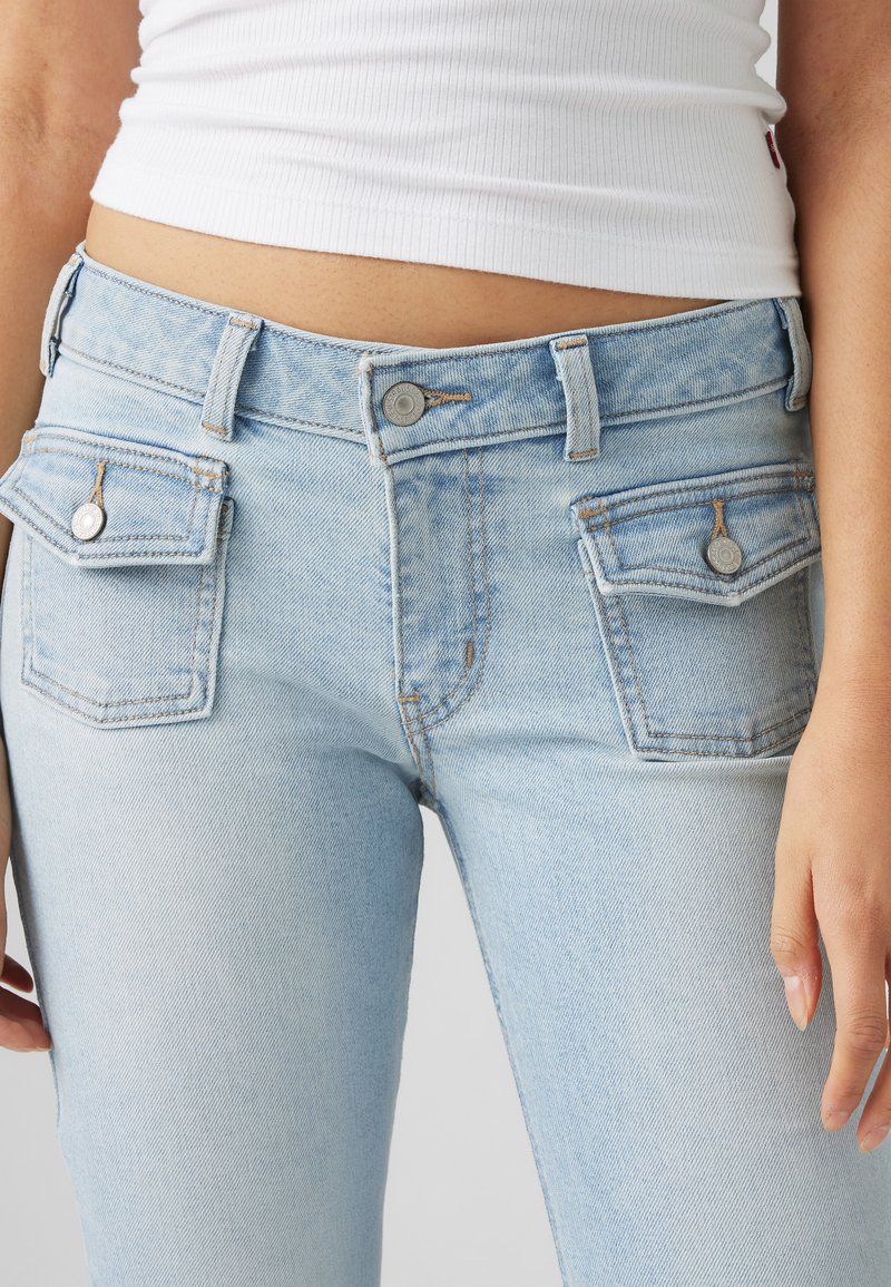 Jean en denim bleu clair avec un design ajusté, deux poches avant avec des accents de bouton, et une fermeture éclair. Le tissu a une texture douce et usée.