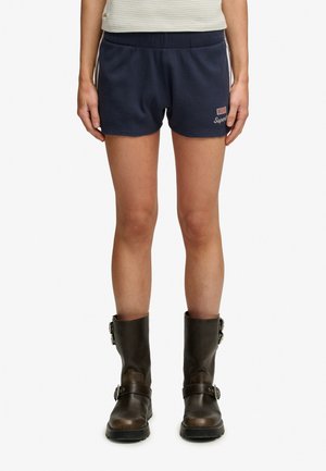 Person iført marineblå shorts med et amerikansk flagmærke og mørkebrune støvler med spænde, stående mod en ensfarvet baggrund.