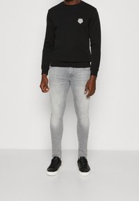 Svart sweatshirt med en liten vit logotyp på bröstet, i kombination med ljusgrå slim-fit jeans och svarta sneakers med vita sulor.