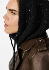 Zwarte gebreide capuchon met pailletten, gedragen met een bruine leren jas. De capuchon heeft een textuurdesign en strikjes. Het zijaanzicht toont de gelaatstrekken.