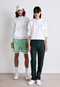 Du modeli stovi šalia vienas kito, vilkėdami baltus džemperius su Lacoste logotipais; vienas apsirengęs žaliais raštuotais šortais ir kepure, kitas - tamsiomis kelnėmis ir akinių nuo saulės.