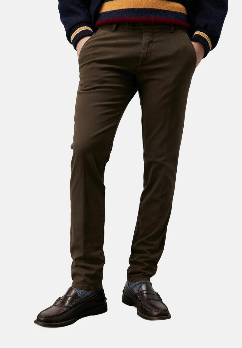 Chinos marroni slim fit con una texture liscia, tasche laterali e chiusura con bottone, abbinati a scarpe scure e calze a contrasto.