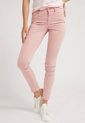 Kobieta w dopasowanych jasnoróżowych dżinsach i białych trampkach, stojąca z jedną nogą lekko ugiętą na tle jednolitego tła.