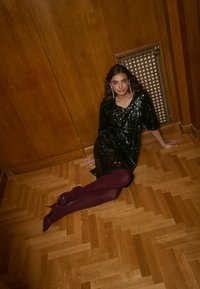 Fekete flitteres ruha laza szabással, bordó harisnyával és pántos magassarkúval párosítva. A háttérben barna, átlós mintázatú fa padló.