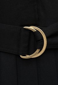 Μαύρη ζώνη υφάσματος με στρογγυλή χρυσή αγκράφα που φέρει χαραγμένη επωνυμία. Η ταινία έχει υφή, προσδίδοντας δομημένη εμφάνιση.