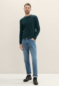 Pull en maille verte à col rond, associé à un jean bleu clair et des chaussures noires à lacets, debout sur un fond neutre.