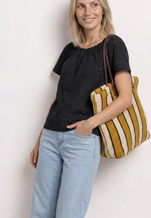 Femme portant un chemisier noir et un jean bleu clair, portant sur une épaule un grand sac cabas rayé tricoté, souriante et regardant ailleurs.