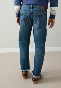 Dżinsy w średnim niebieskim kolorze o prostym kroju nogawek, z tylnymi kieszeniami i podwiniętymi mankietami. Do stroju dopasowane brązowe skórzane buty.