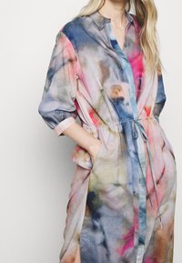 Kobieta ubrana w długą, kolorową sukienkę tie-dye z podwiniętymi rękawami i paskiem w talii, z rękami w kieszeniach, na jednolitym jasnym tle.