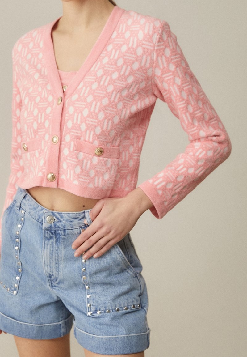 Różowy skrócony kardigan z teksturowanym geometrycznym wzorem, z złotymi guzikami i dwiema przednimi kieszeniami, zestawiony z jasnoniebieskimi szortami dżinsowymi.