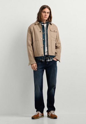 Jonge man met schouderlang haar die een beige jas draagt over spijker- en geruite hemden, donkere jeans en bruine sneakers, staand tegen een witte muur.