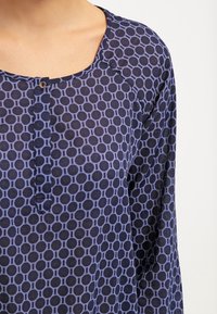 Långärmad blus i marinblått med ett cirkulärt mönster i ljusare nyanser. Den har en rund krage med en enda knappstängning.