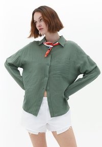 Camicia verde con bottoni, dal finish testurizzato, con tasca sul petto e vestibilità ampia. Indossata sopra pantaloni corti bianchi strappati e accessoriata con una sciarpa a fantasia.