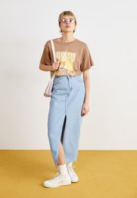 Bruine graphic t-shirt met korte mouwen, lichtblauwe denim rok met een voorkniplijn, witte hoge sneakers en lichtroze zonnebril.