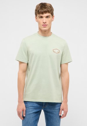 Junger Mann trägt ein hellgrünes T-Shirt mit kurzen Ärmeln und kleinem "Mustang"-Logo auf der linken Brust, kombiniert mit blauen Jeans.