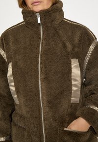 Flísová bunda v tmavohnedej farbe s vysokým golierom, striebornými saténovými detailmi na bokoch a rukávoch a predným zipsom. Má bočné vrecká.