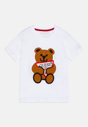 Hvid t-shirt med korte ærmer med en brun tøjbjørn, der holder et rød-hvidt trekantet flag med "GUESS U.S.A." trykt på.