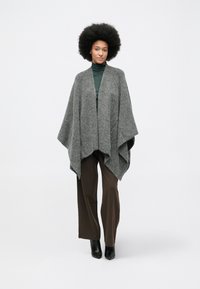 Grå, strikket poncho med en løs, drapert design. Båret over en mørkegrønn turtleneck og kombinert med brune bukser og svarte hælbukser.