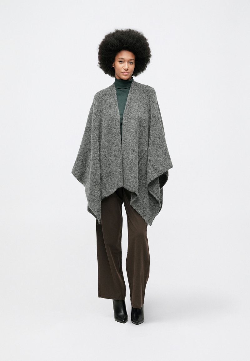 Grå, strikket poncho med en løs, drapert design. Båret over en mørkegrønn turtleneck og kombinert med brune bukser og svarte hælbukser.