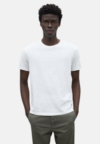 Camiseta blanca de algodón con cuello redondo y mangas cortas, luciendo en un modelo que lleva pantalones verde oliva, con un fondo neutro.