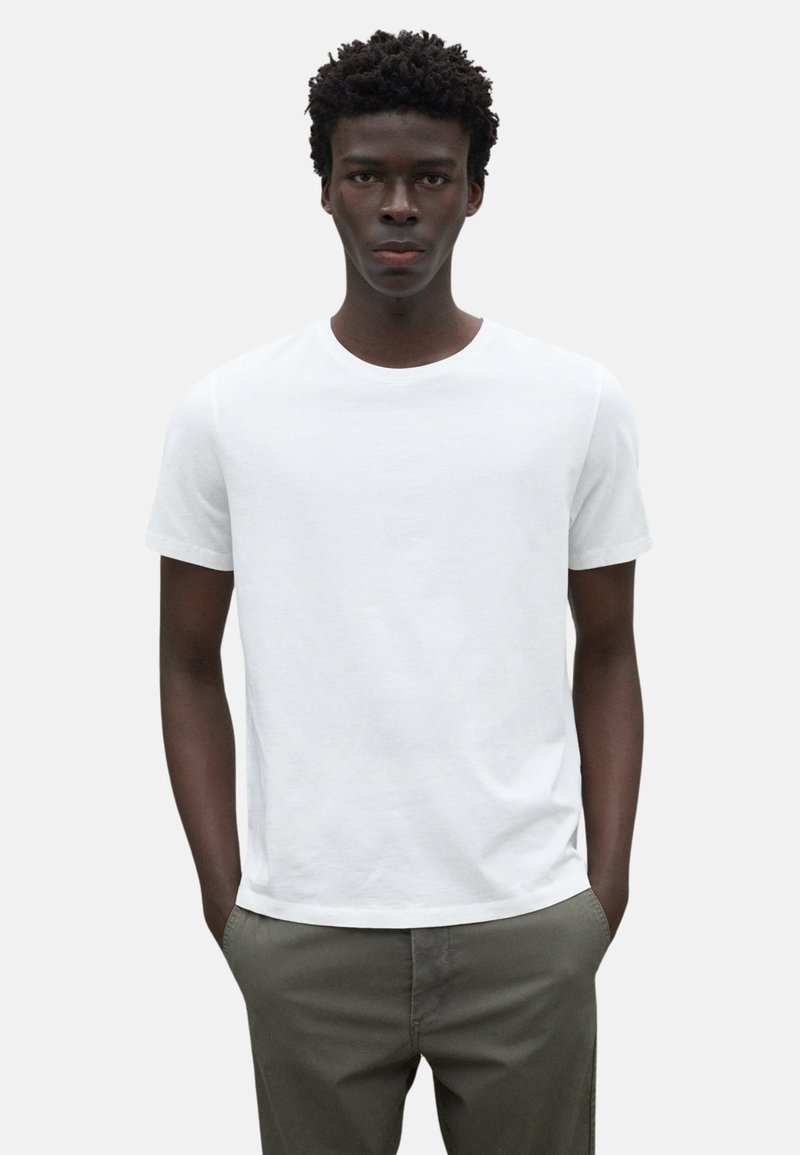 Camiseta blanca de algodón con cuello redondo y mangas cortas, luciendo en un modelo que lleva pantalones verde oliva, con un fondo neutro.