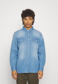 Camisa de mezclilla azul de botones con dos bolsillos en el pecho, un cuello clásico y mangas largas con un ajuste relajado. Tejido texturizado, desvanecido sutilmente.