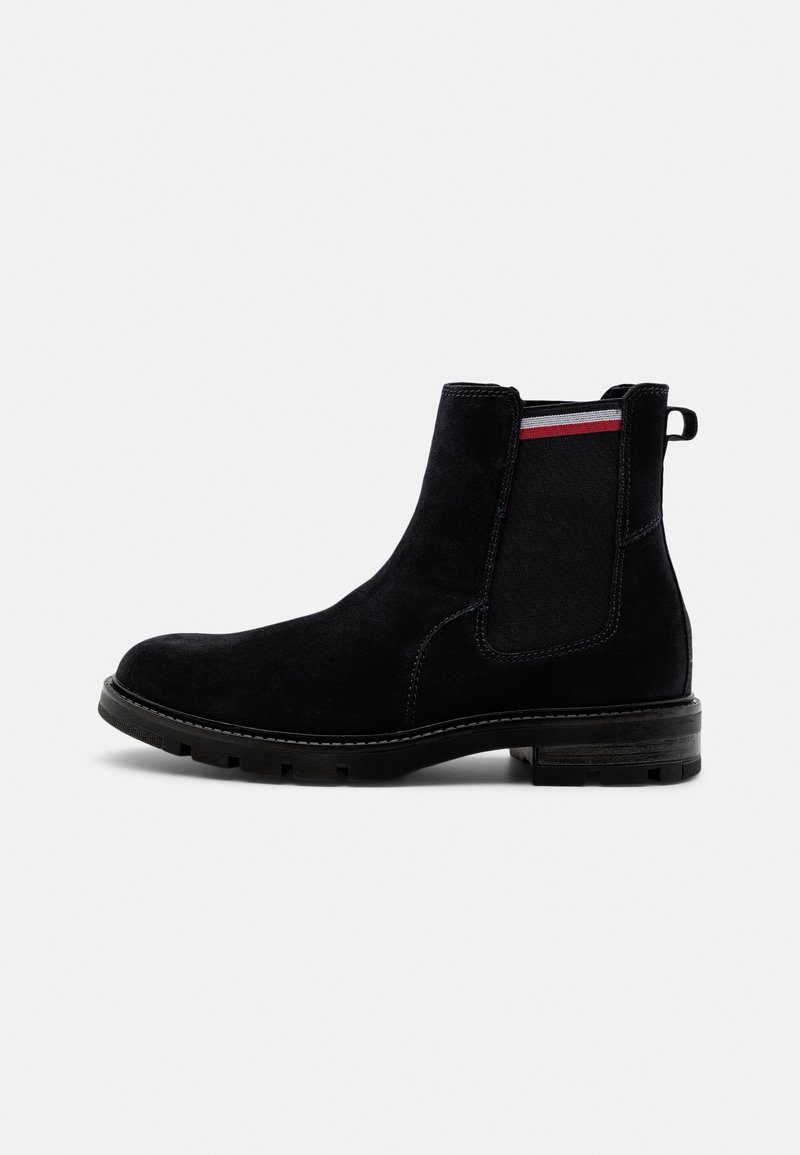 Botte Chelsea en daim noir avec semelle en caoutchouc, panneau latéral élastique avec bande rouge et blanche, et languette à l'arrière.