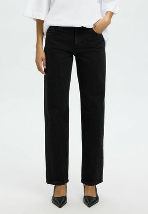 Kobieta ubrana w czarne proste jeansy, biały top z długim rękawem i czarne buty na obcasie z ostrym noskiem, stoi z jedną ręką w kieszeni.