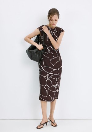 Femme en robe midi fleurie noire et blanche ajustant un grand sac cabas noir, portant des sandales à talons noires, debout contre un mur blanc.