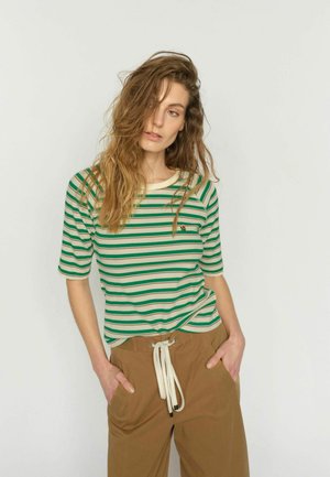 Naine, kellel on pikad lainelised juuksed, kannab rohelist ja kreemjat triibulist lühikeste varrukatega pluusi ning pruune kõrge vöökohaga pükse valgete nööridega, käed taskutes.