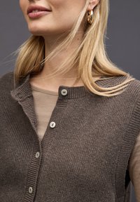Gilet en tricot côtelé marron avec un col rond, doté de deux boutons clairs et superposé à un haut beige. Texture lisse, design moderne.