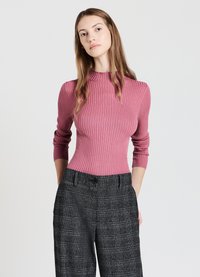 Enganliegender gerippter Rollkragenpullover in staubigem Rosa, lange Ärmel mit umgeschlagenen Details, kombiniert mit hoch taillierten grauen Karottenhosen mit Knopfverschluss.